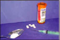 Photograph showing a spoon holding a white powder, next to a syringe, next to several white tablets lying beside a pill bottle.
