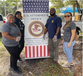 Community Outreach team at an outdoor event, posing next to sign with the USAO-SDFL seal.