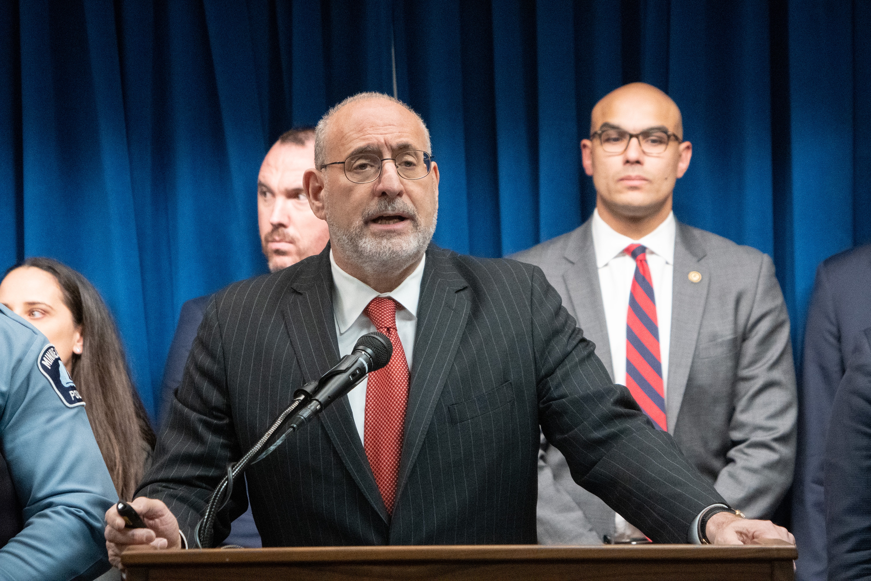 United States Attorney for the District of Minnesota Andrew M. Luger delivers remarks from a podium at the United States Attorney’s Office for the District of Minnesota office in Minneapolis, Minnesota. Other officials stand in a row behind.