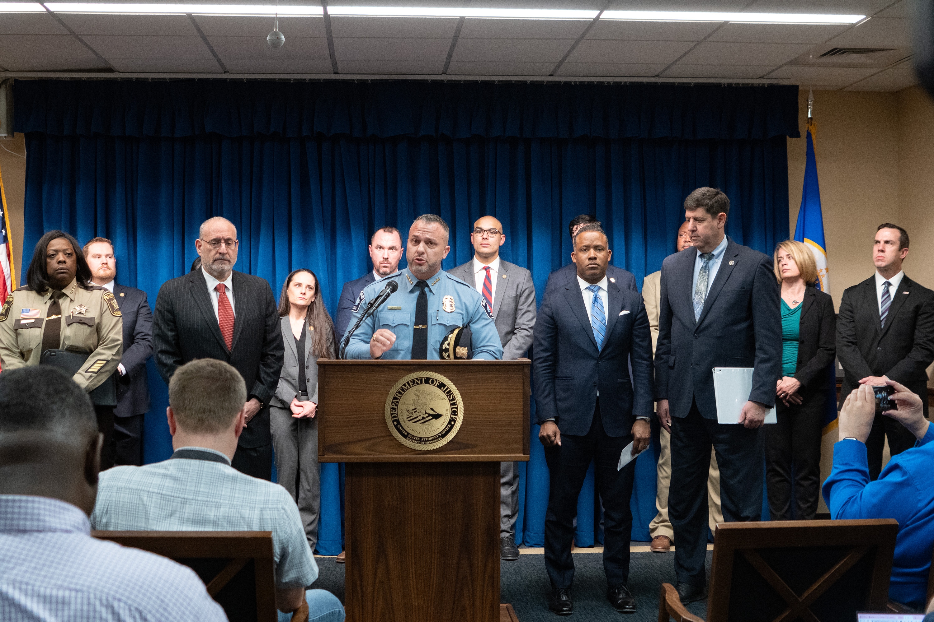 Chief of the Minneapolis Police Department Brian O'Hara delivers remarks from a podium bearing the United States Attorney's Office for the District of Minnesota seal. To the left stands United States Attorney for the District of Minnesota Andrew M. Luger (left) and Hennepin County Sheriff Dawanna S. Witt (far left). To the right stands Assistant Attorney General Kenneth A. Polite, Jr. (right) and ATF Director Steven M. Dettelbach (far right). Other officials stand in a row behind.