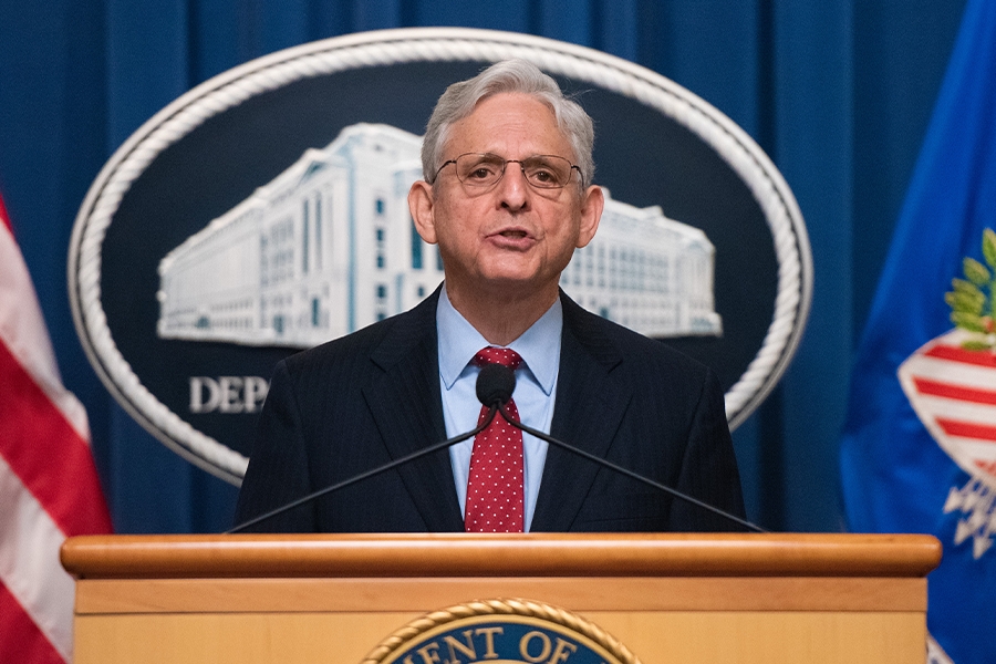 Attorney General Merrick B. Garland delivers remarks on the podium. The Department of Justice seals are in front of and behind him. The DOJ blue flag is on the right, and the red and white stripes of the American flag are to the left.