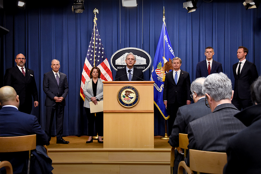 Merrick B. Garland speaks at the podium. To the left are Assistant Attorney General Matthew Olsen and Deputy Attorney General Lisa O. Monaco. To the right of the Attorney General is Christopher Wray, Director of the Federal Bureau of Investigation (FBI). An American flag and the Department of Justice flag in blue are seen in the background.