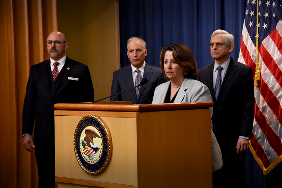 Deputy Attorney General Lisa O. Monaco speaks at a podium. On her left is Assistant Attorney General Matthew Olsen, and to the right is the Attorney General Merrick B. Garland.