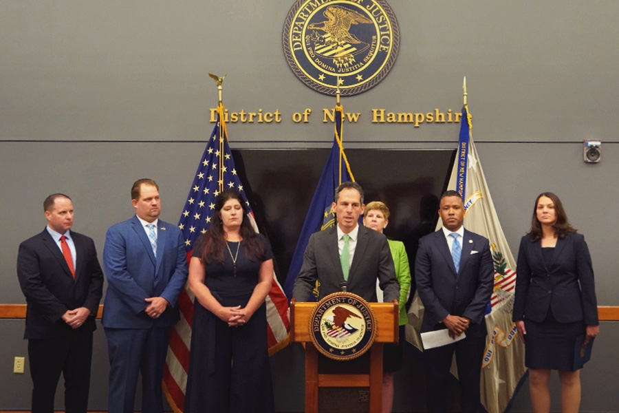 U.S. Attorney Nikolas P. Kerest for the District of Vermont speaks at the podium with a Department of Justice seal. The background displays the seal of the district of New Hampshire.