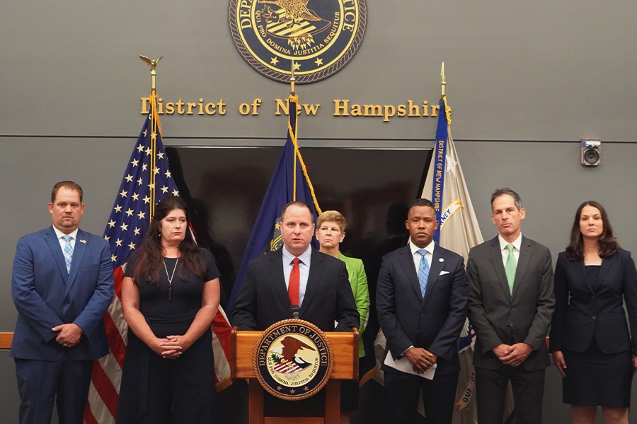 Acting Deputy Assistant Director Aaron Tapp of the FBI's Criminal Investigative Division speaks at the podium with a Department of Justice seal. The background displays the seal of the district of New Hampshire.