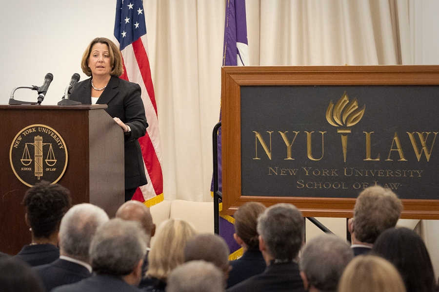 Deputy Attorney General Lisa O. Monaco speaks to an audience of officials and attorneys at a podium bearing the New York University seal. To the right is a blackboard that says, “NYU LAW, New York University School of Law”