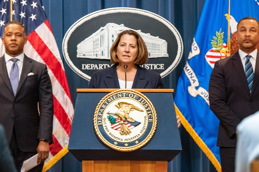Deputy Attorney General Lisa O. Monaco delivers remarks from a podium bearing the Department of Justice seal. To the left stands Assistant Attorney General Kenneth A. Polite Jr. of the Justice Department's Criminal Division. To the right stands U.S. Attorney Breon Peace for the Eastern District of New York.