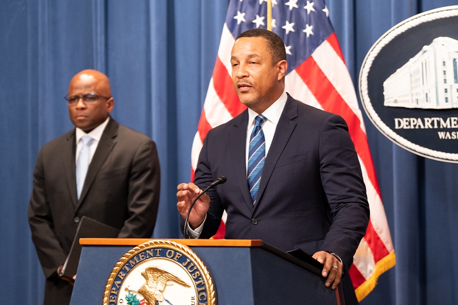 U.S. Attorney Breon Peace for the Eastern District of New York delivers remarks from a podium bearing the Department of Justice seal. To the left stands Associate Deputy Director Brian Turner of the FBI.
