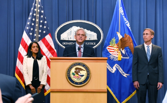 Attorney General Merrick B. Garland speaks at a podium bearing the Department of Justice seal. To the left is Associate Attorney General Vanita Gupta, who stands in front of the American flag. To the right stands the Principal Deputy Assistant Attorney for the Civil Division Brian Boynton.