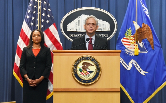 Attorney General Merrick B. Garland speaks at a podium bearing the Department of Justice seal. To the left is Assistant Attorney General Kristen Clarke, who stands in front of the American flag.