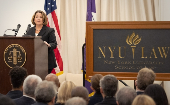 Deputy Attorney General Lisa O. Monaco speaks to an audience of officials and attorneys at a podium bearing the New York University seal. To the right is a blackboard that says, “NYU LAW, New York University School of Law”