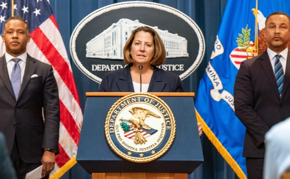 Deputy Attorney General Lisa O. Monaco delivers remarks from a podium bearing the Department of Justice seal. To the left stands Assistant Attorney General Kenneth A. Polite Jr. of the Justice Department's Criminal Division. To the right stands U.S. Attorney Breon Peace for the Eastern District of New York.