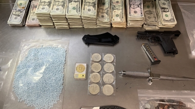 picture of evidence on a table. Evidence includes, gun, cash, knives, coins, blue pills, and other dangerous weapons