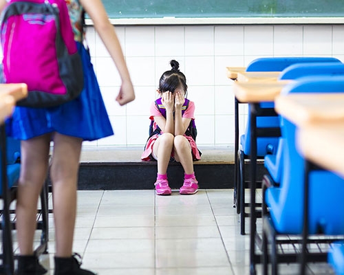 A young Asian American student covers her hands over her eyes as other students look on. 
