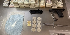 picture of evidence on a table. Evidence includes, gun, cash, knives, coins, blue pills, and other dangerous weapons